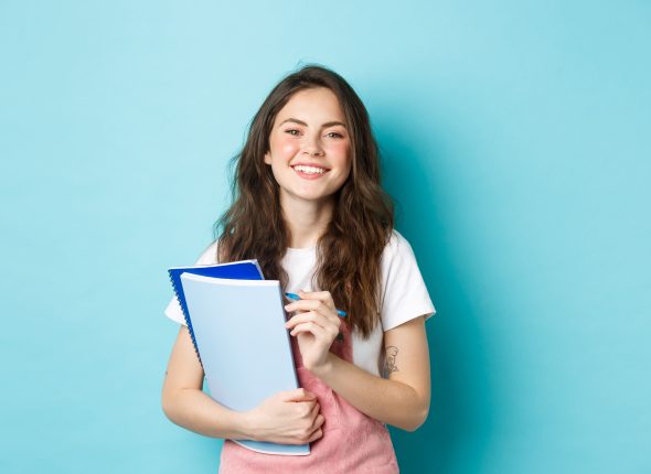 happy-young-female-student-holding-notebooks-from-courses-and-smiling-at-camera-standing-in-spring-clothes-against-blue-background-1-scaled